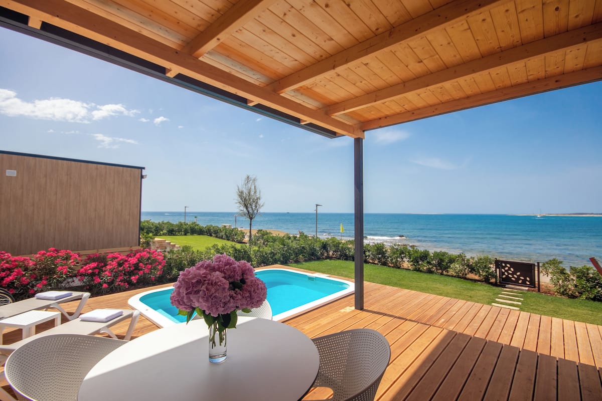 A wooden deck with a pool, lounge chairs, and a table with a vase of flowers.