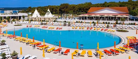 A large swimming pool surrounded by sun loungers and umbrellas, with a building and trees in the background.