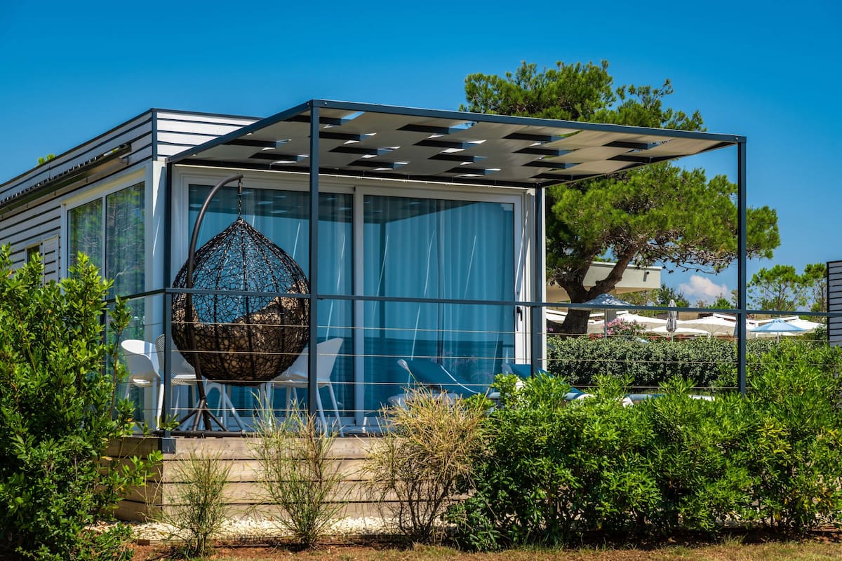 A modern glass house with a hanging chair, surrounded by greenery and a clear sky.