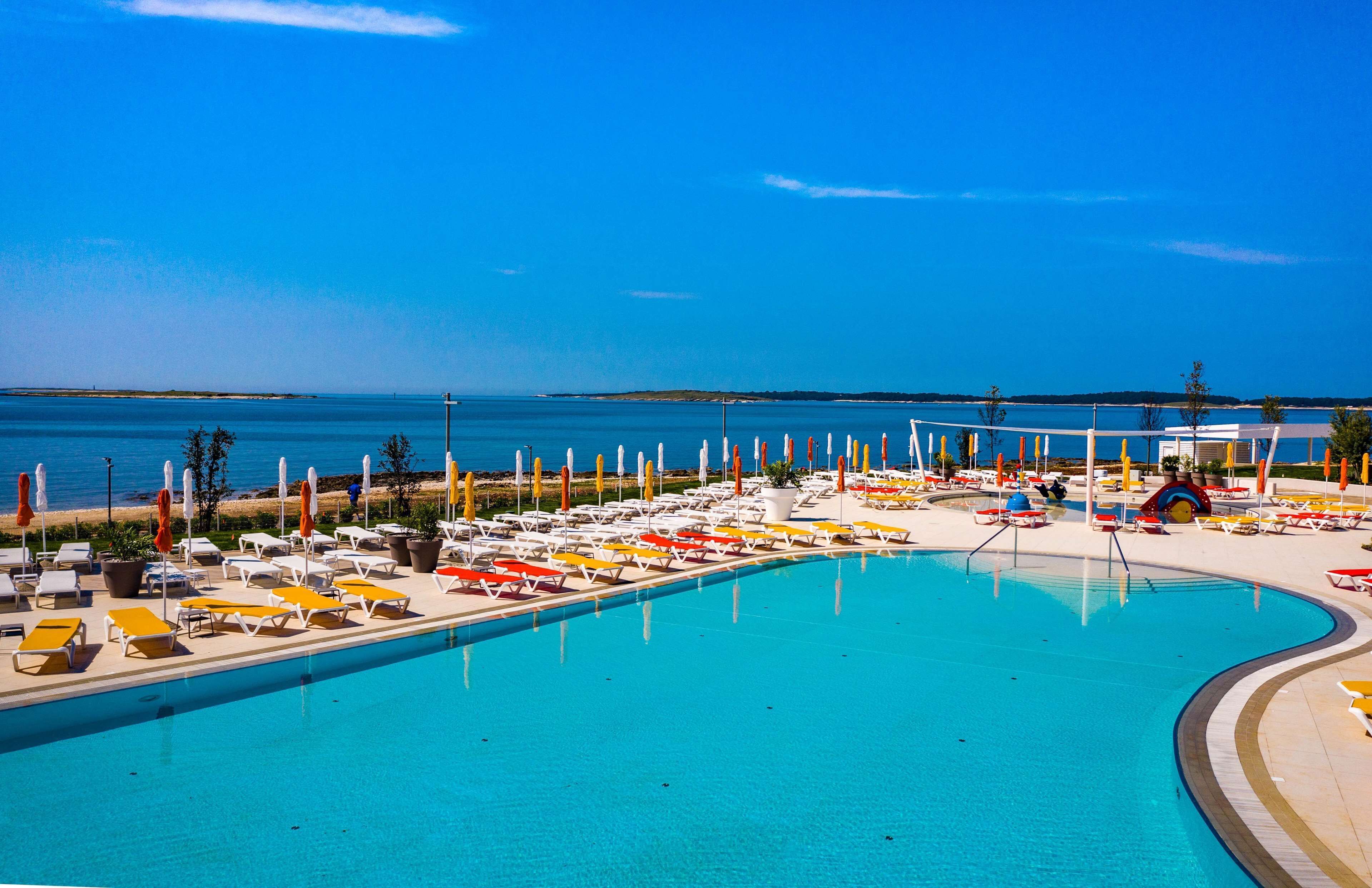 Une grande piscine avec des chaises longues et des parasols, entourée d’une plage et offrant une vue sur l’océan.