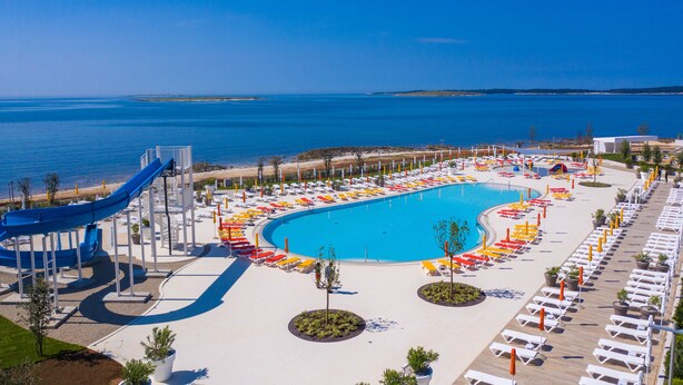 En stor swimmingpool med en blå vattenrutschkana, omgiven av solstolar och parasoller, med en strand och havet i bakgrunden.