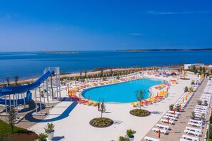 A large swimming pool with a blue slide, surrounded by sun loungers and umbrellas, with a beach and ocean in the background. - Arena Grand Kazela Camping Homes (Medulin)