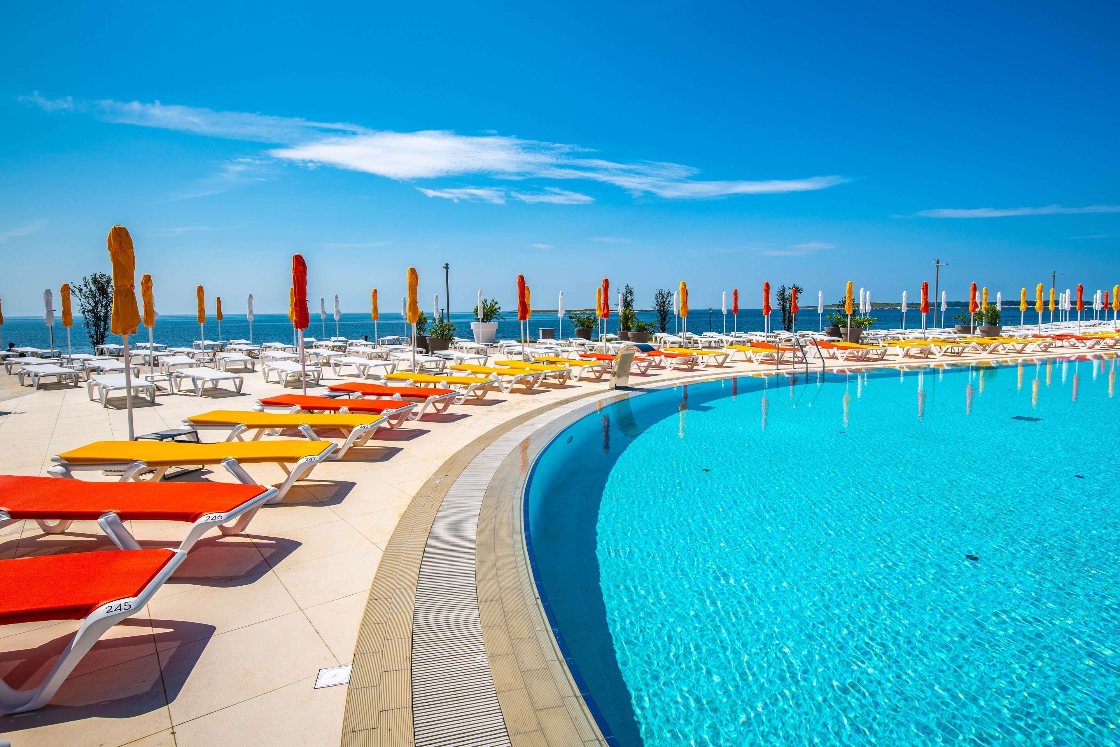 Ein Poolbereich mit Liegestühlen und Sonnenschirmen, ein klarer, blauer Himmel und eine ruhige See im Hintergrund.