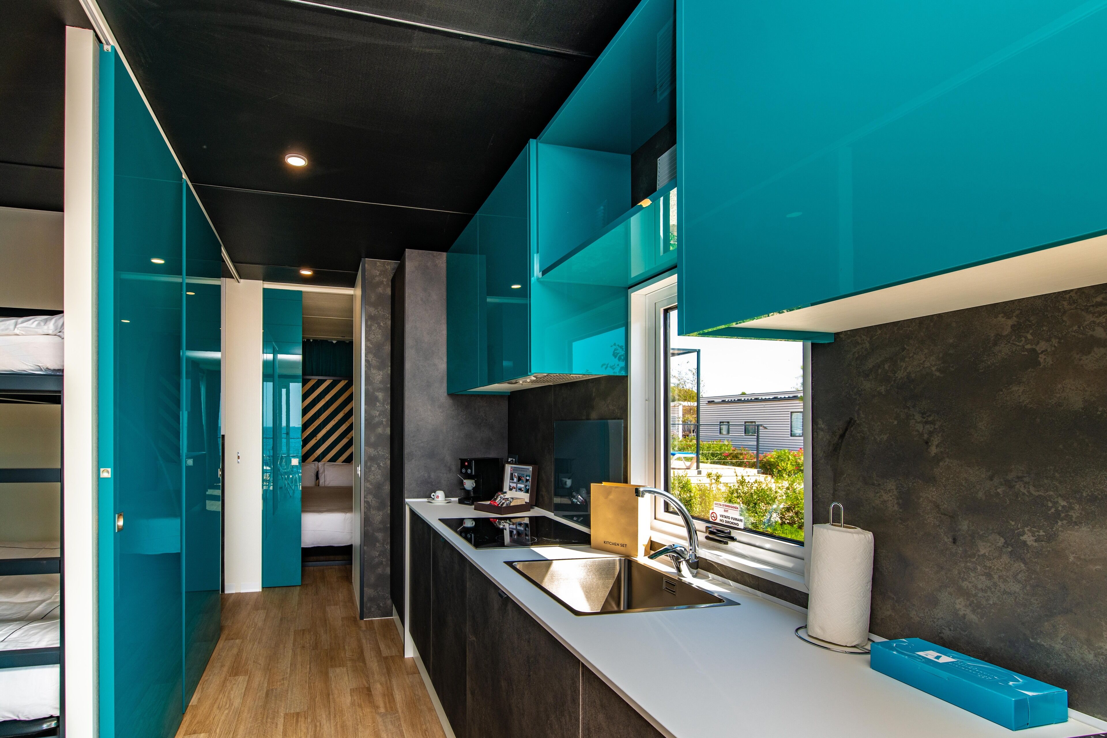 Eine moderne Küche mit türkis-blauen Schränken, Holzboden und einem großen Fenster mit Blick nach draußen.