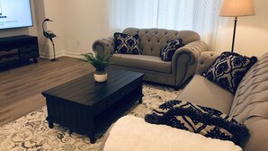 Ein Wohnzimmer mit einem dunklen Couchtisch, zwei Sofas mit gemusterten Kissen, einer Zimmerpflanze und einem Fernseher an der Wand.