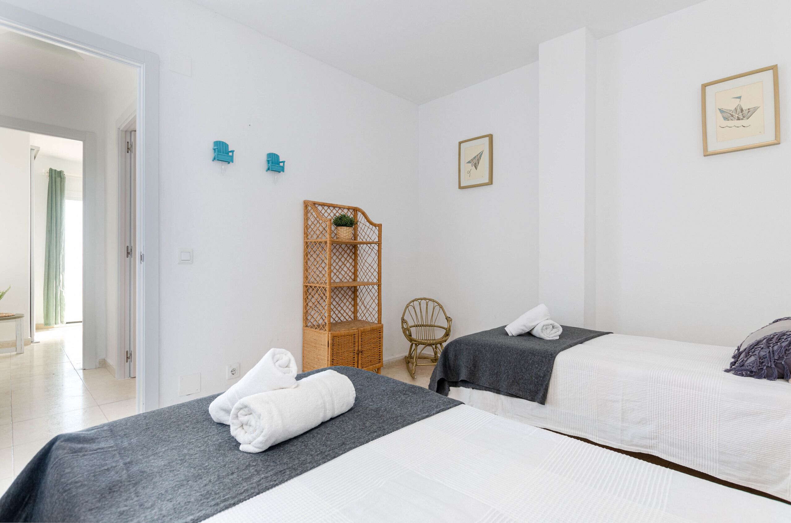 A hotel room with two beds, a wicker chair, a small table, and a door leading to a bathroom.