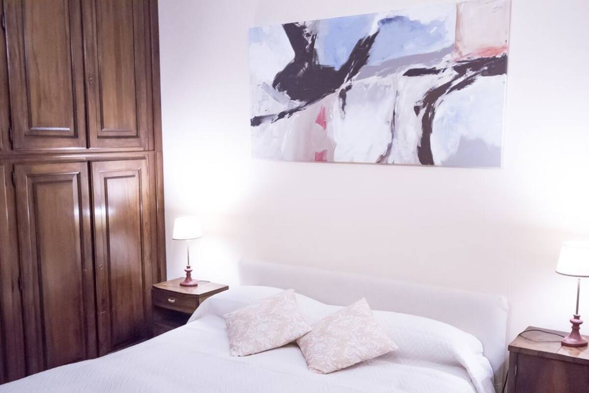 A hotel room with a large bed, two bedside tables with lamps, a wooden wardrobe, and a modern abstract painting on the wall.