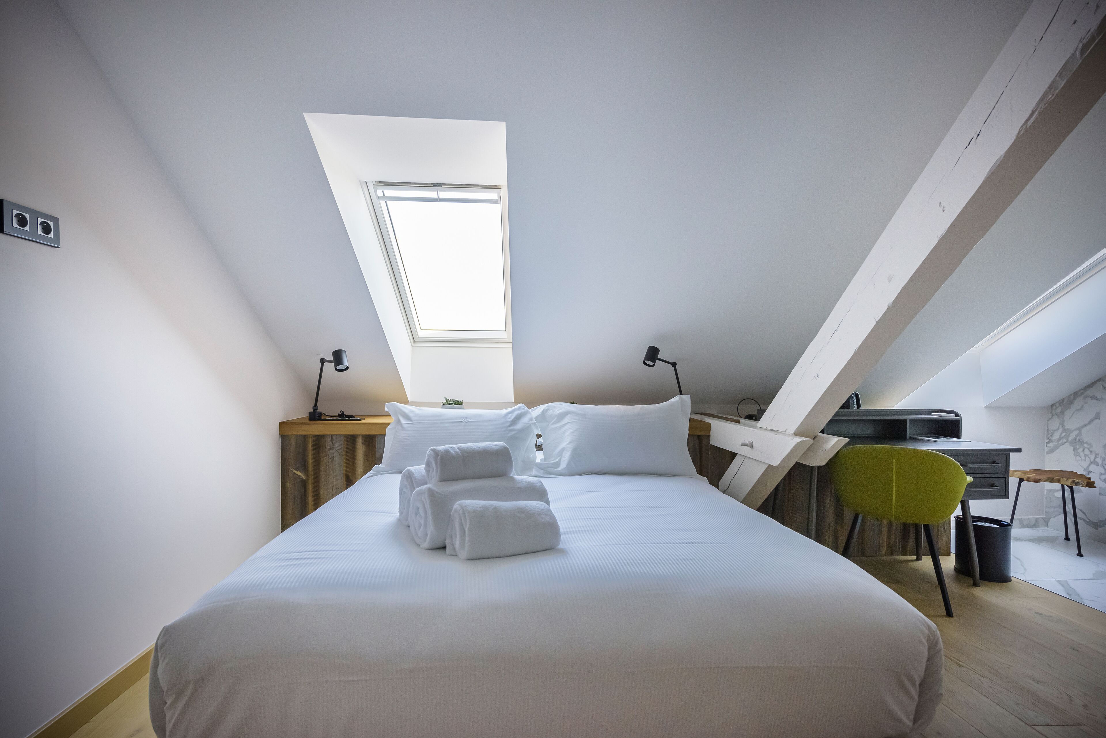 A modern bedroom with a skylight, a bed with neatly folded towels, a desk, and a bright green chair.