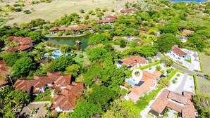 Vista aérea de un complejo turístico costero con varias construcciones, un cuerpo de agua central y abundante vegetación.