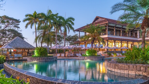 Un resort con piscina, sillones de descanso y un restaurante con sombrillas exteriores.