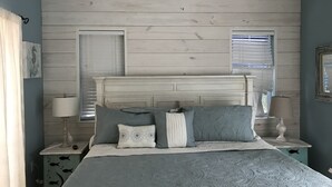 Ein Schlafzimmer mit einem Holzkopfteil, einem Bett mit gemusterten Betttextilien, zwei Nachttischen mit Lampen und einem Fenster mit Jalousien.