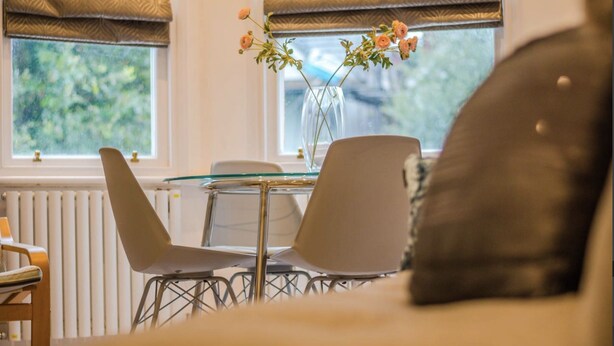 A room with a dining area featuring a glass table and metal chairs, a vase with flowers on the windowsill, and a view of greenery outside.