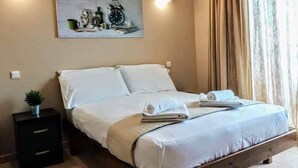 A neatly made bed with white linens and a folded towel, a small bedside table with a plant, and a painting of clocks on the wall. - Dhermi Lighthouse (Dhërmi)