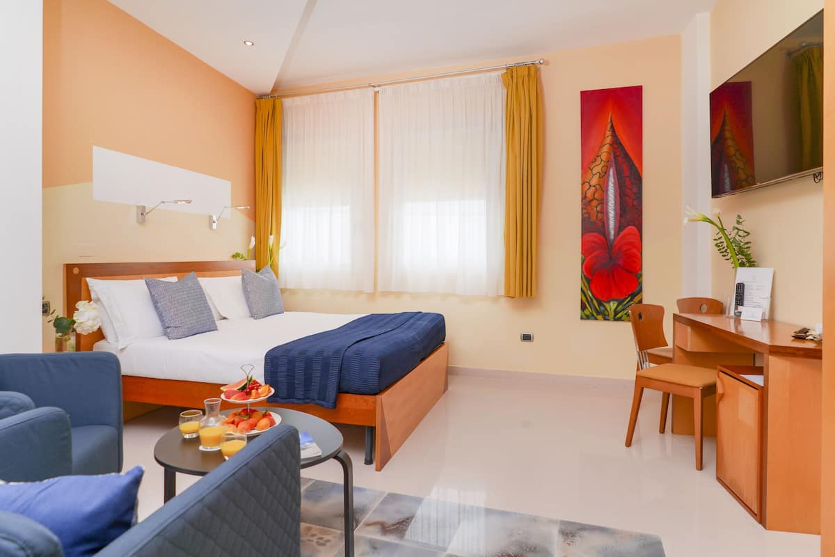 A hotel room with a bed, a desk, a chair, and a small table with fruit.