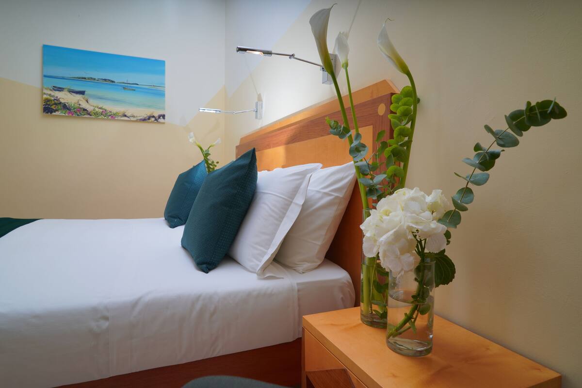 A hotel room with a bed, pillows, a nightstand with a vase of flowers, a painting on the wall, and a lamp.
