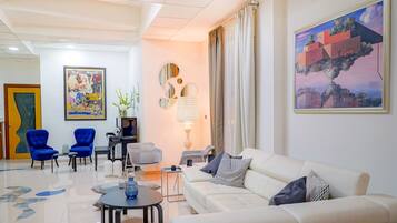 Ein modernes Wohnzimmer mit einem weißen Sofa, blauen Sessel und einem Esstisch. An der Wand hängt ein Gemälde und in der Ecke steht eine Lampe.