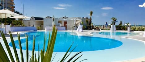 A swimming pool with a wavy edge, surrounded by lounge chairs and umbrellas, with a building in the background.