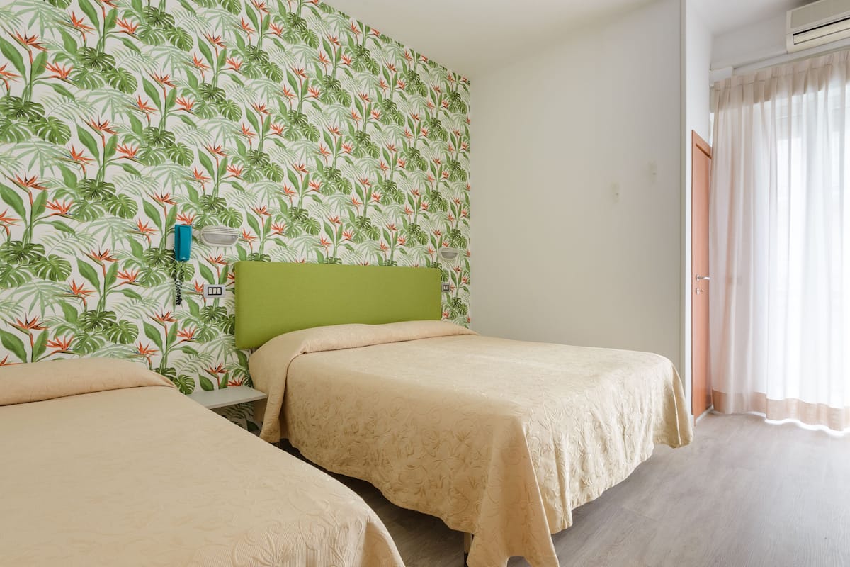 A hotel room with two beds, a green headboard, a floral wallpaper, and a window with curtains.