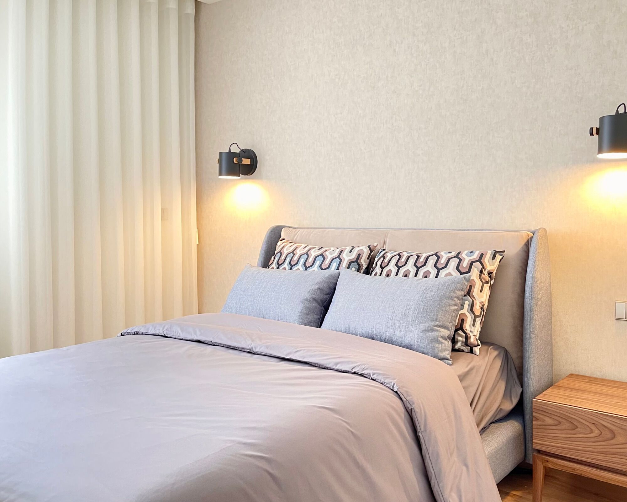 A neatly made bed with a light-colored duvet, two pillows with a geometric pattern, and a wooden nightstand with a lamp.