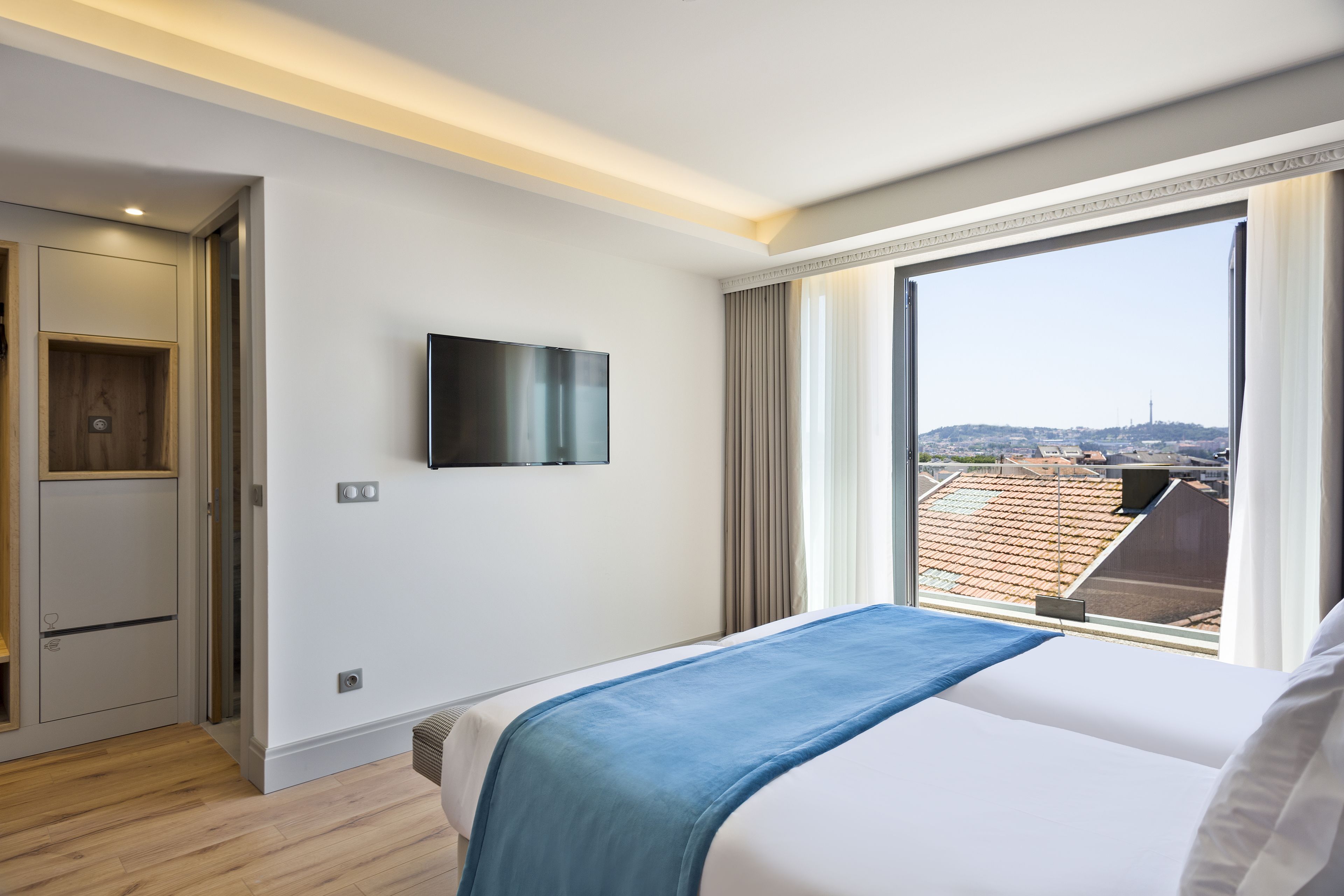 A hotel room with a bed, a television mounted on the wall, and a large window offering a city view.