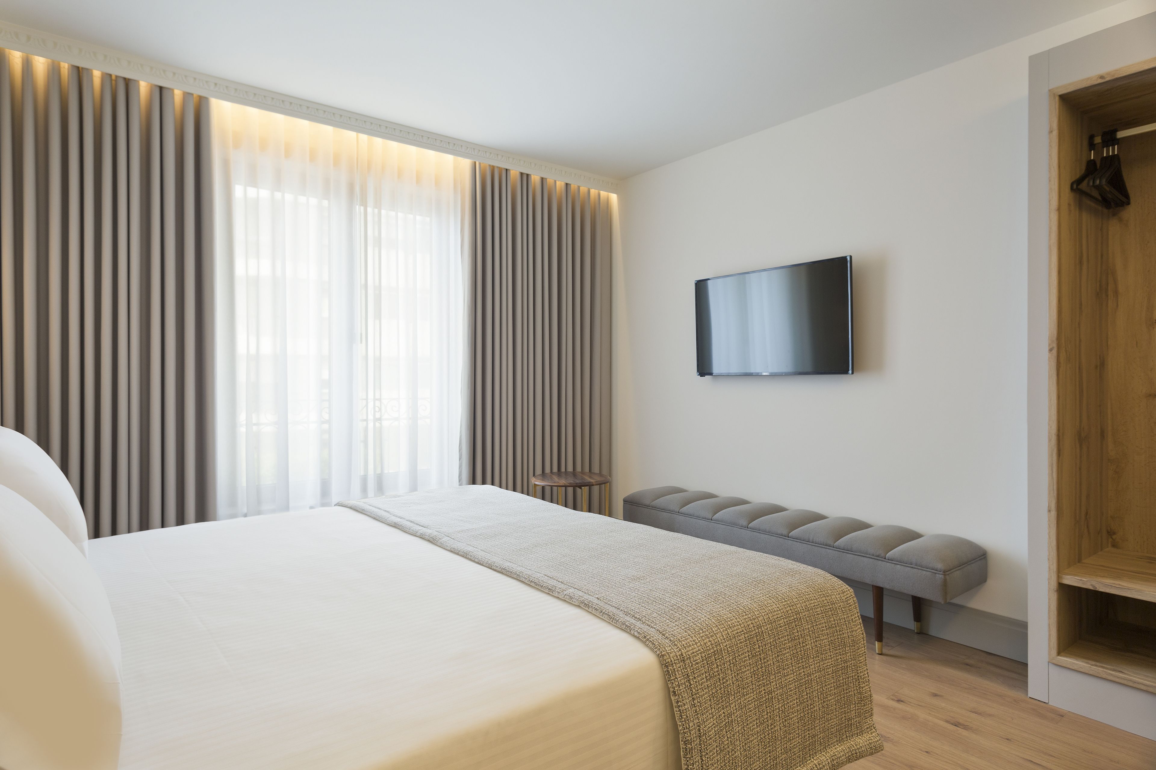 A modern hotel room with a large bed, a flat-screen TV, a bench, and a wooden wardrobe.