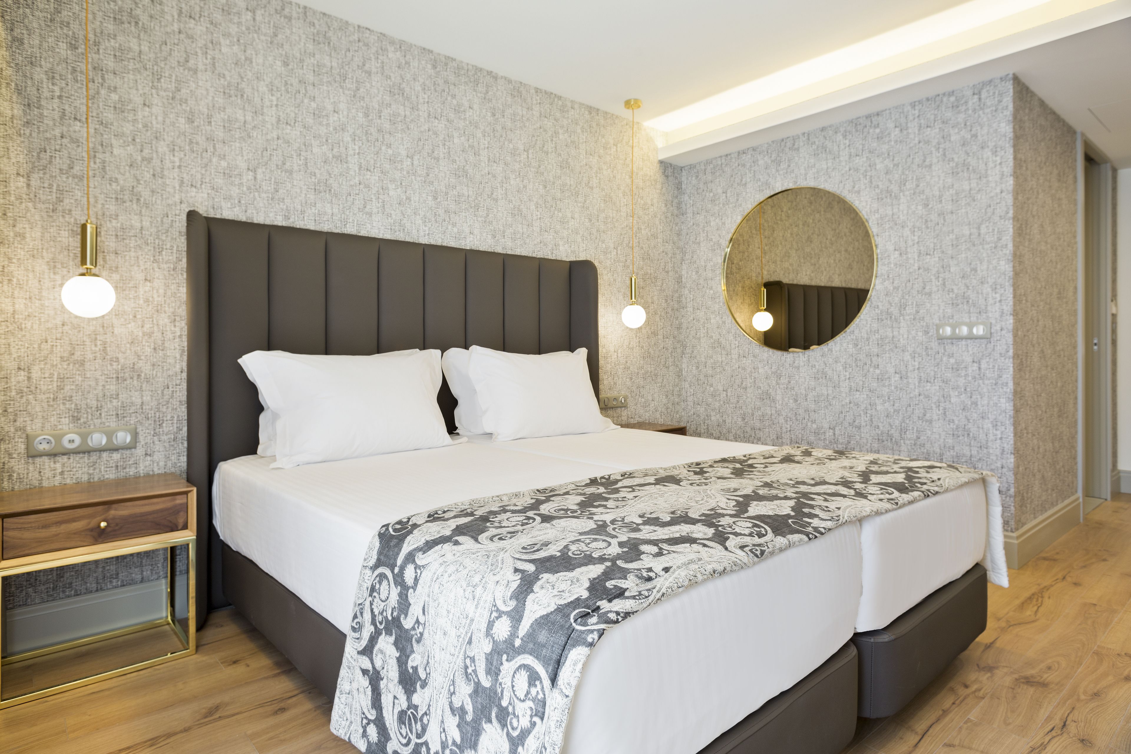 A modern bedroom with a large bed, bedside tables, a round mirror, and a patterned bedspread.