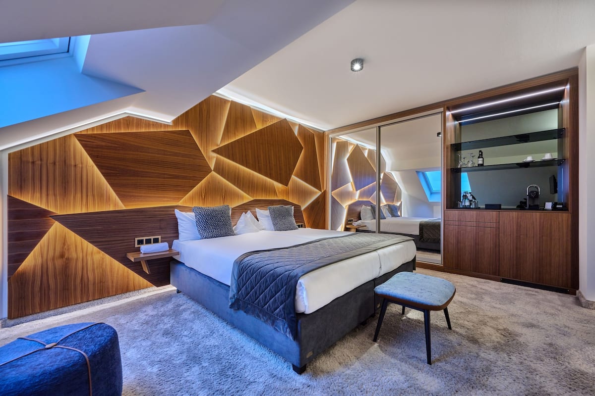 A modern bedroom with a geometric wooden wall design, a large bed, a bench, and a visible kitchenette area.
