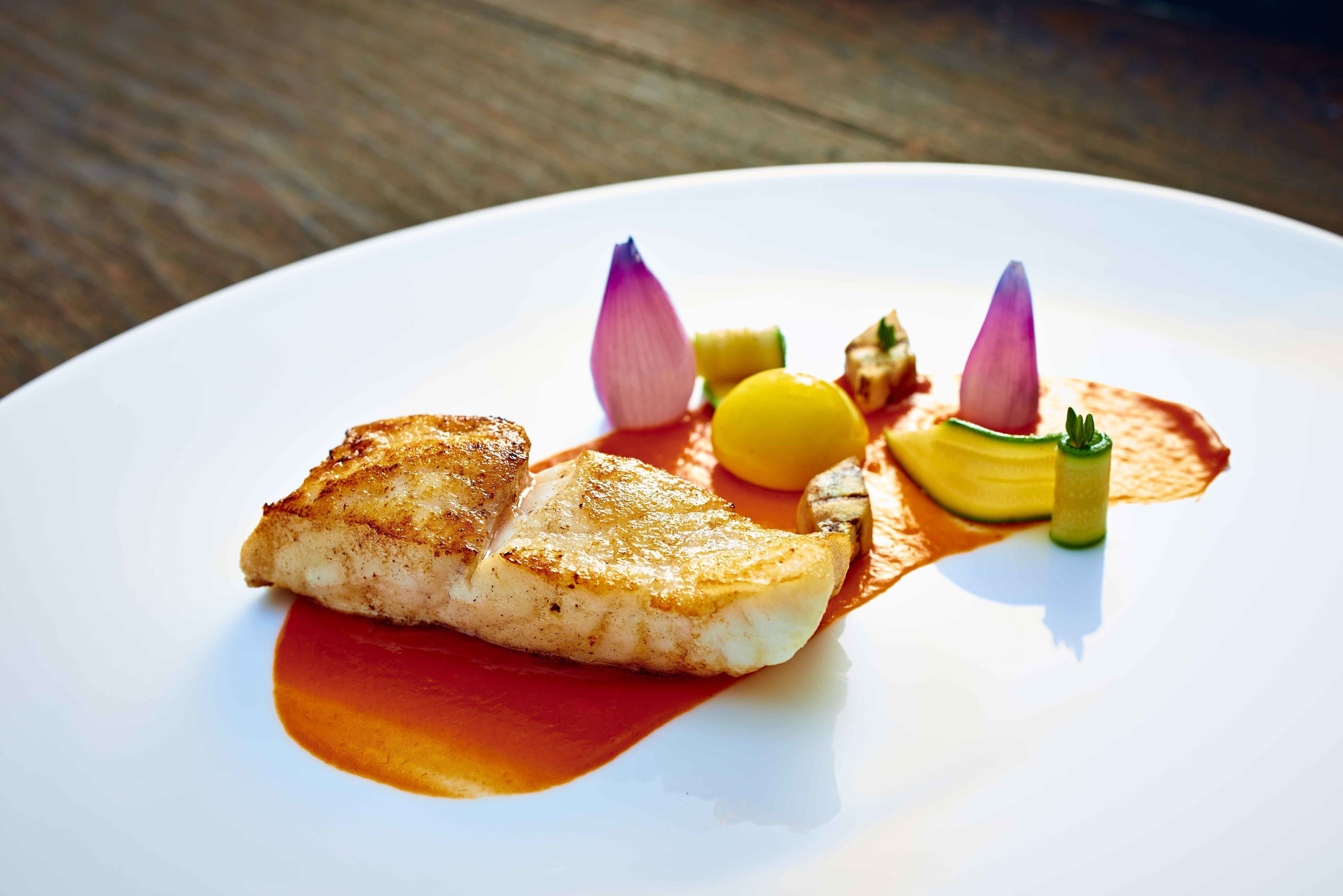 Une assiette blanche contenant un morceau de poisson grillé, une cuillerée de sauce et quelques garnitures.