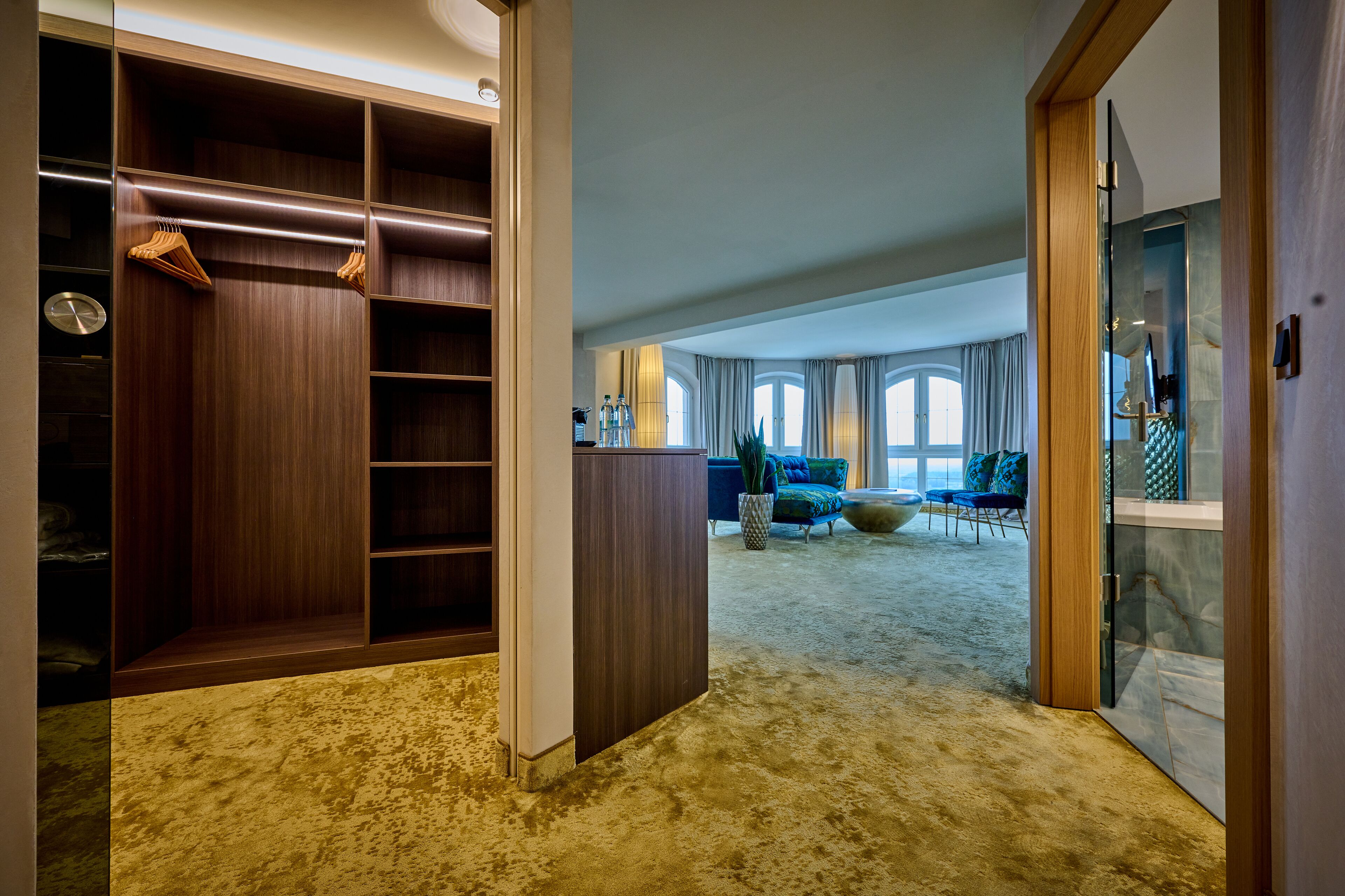 Une pièce spacieuse dotée d’un grand dressing, d’un coin salon avec un canapé et des fauteuils bleus, et offrant une vue sur l’extérieur grâce à de grandes fenêtres.