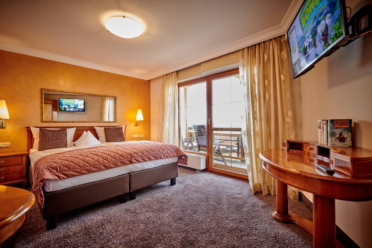 A hotel room with a bed, a TV mounted on the wall, a desk, and a view of the outdoors.