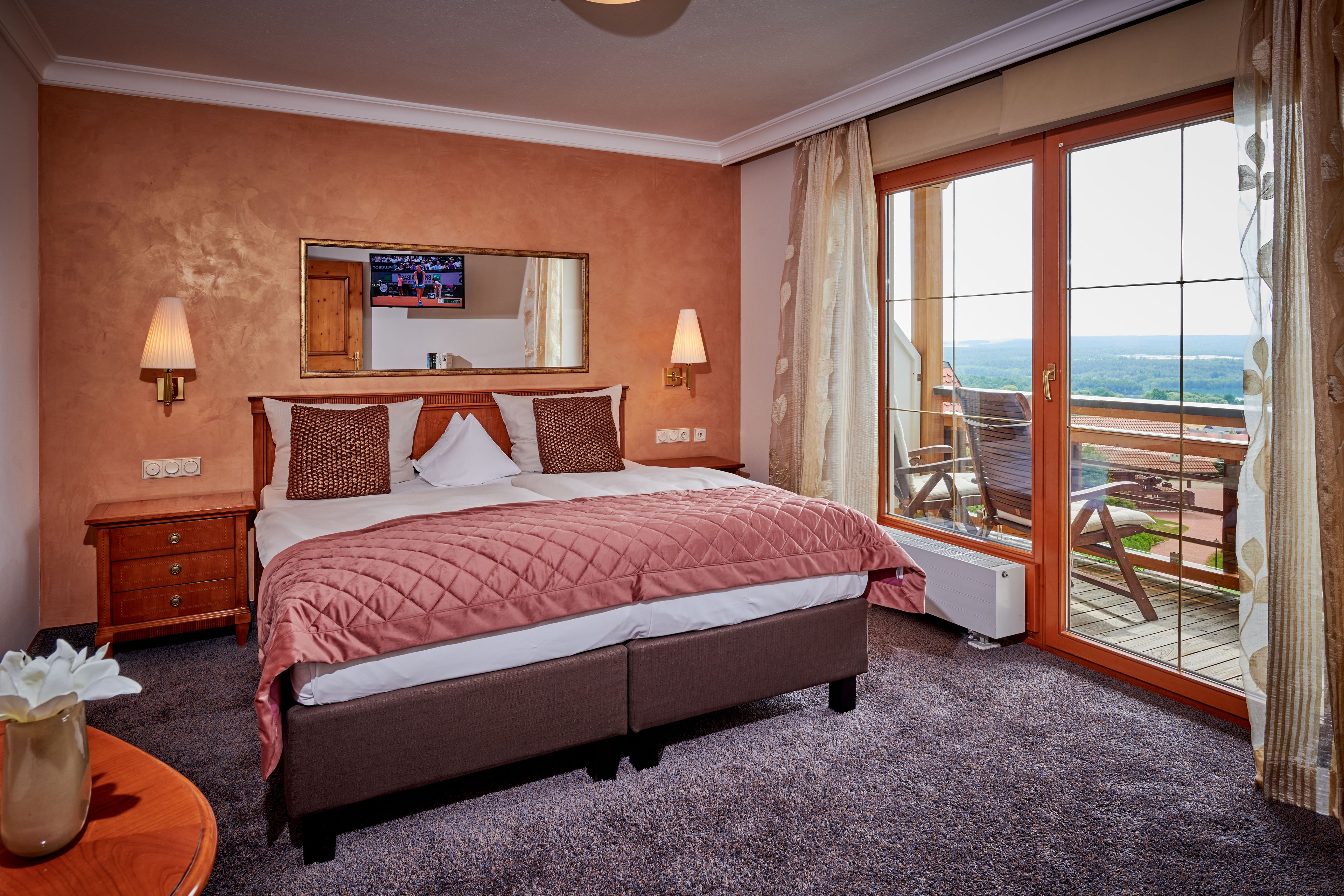 Ein Hotelzimmer mit einem großen Bett, einem Fernseher, einer Glastür zu einem Balkon und einem kleinen Tisch mit einer Vase.