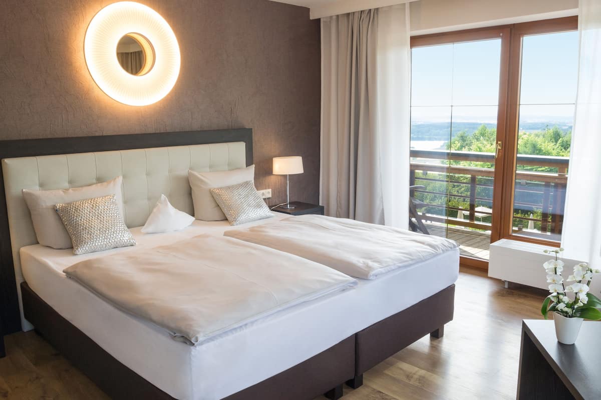 A large bed with multiple pillows, a bedside table with a lamp, a wall-mounted light fixture, and a view of a balcony with a railing.