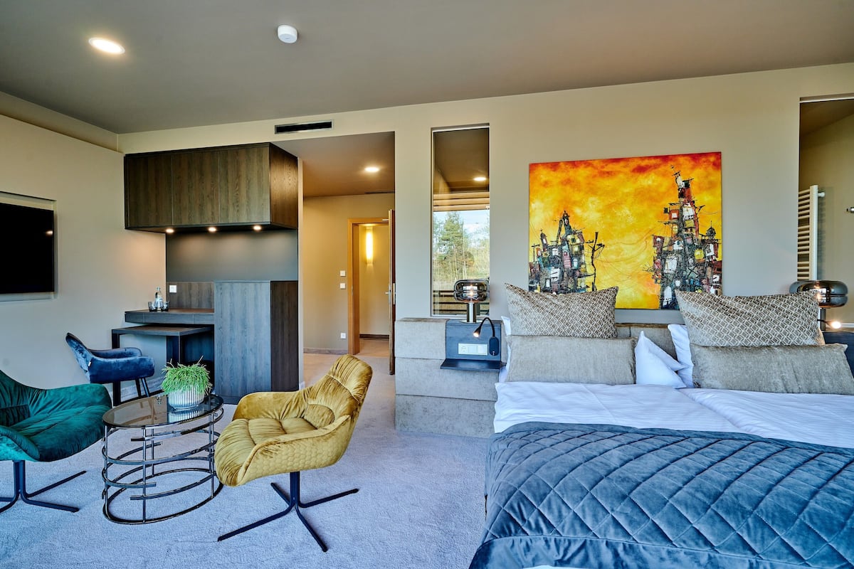 A modern bedroom with a large bed, a desk, and a chair. There is a painting on the wall and a window providing natural light.