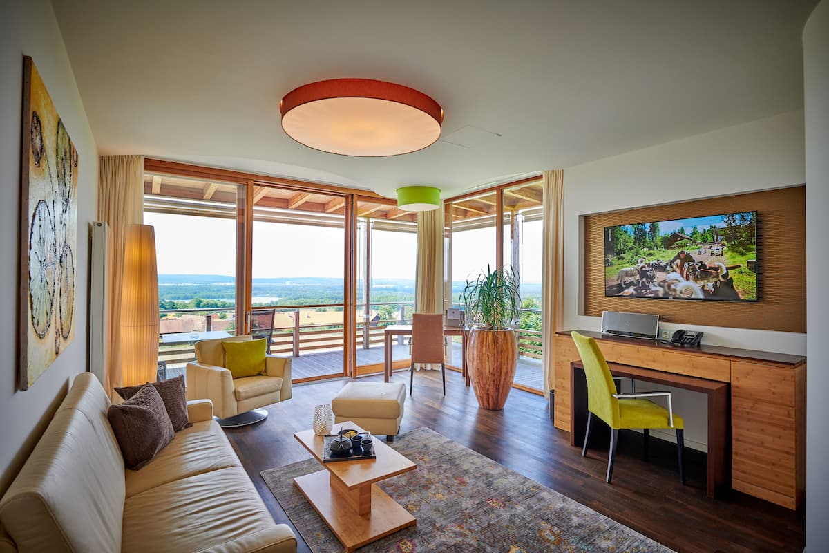 A spacious living area with a sofa, coffee table, and a desk with a chair. There is a large window offering a view of the outdoors.