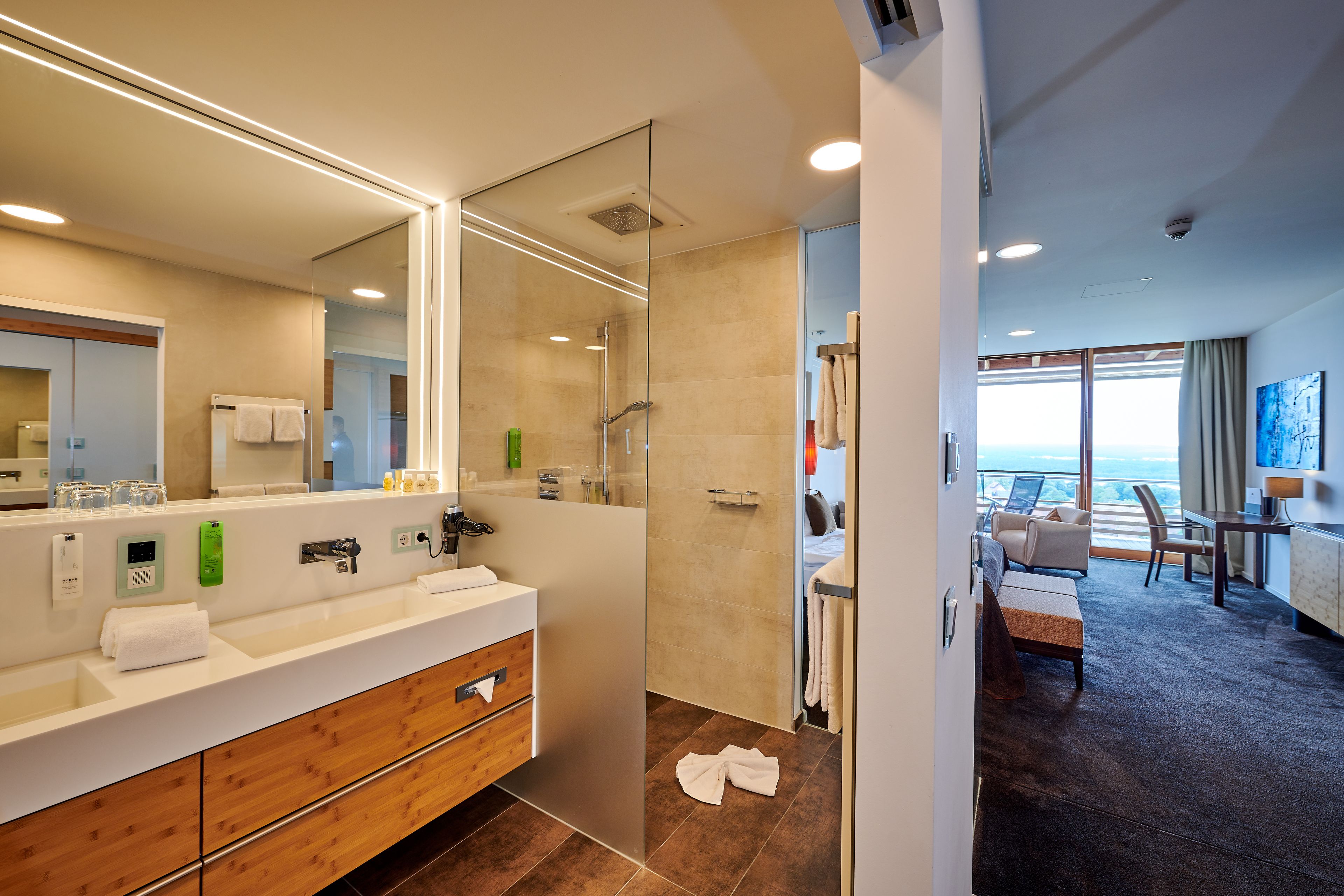 A modern hotel bathroom with a large mirror, a shower, and a vanity with a wooden cabinet.