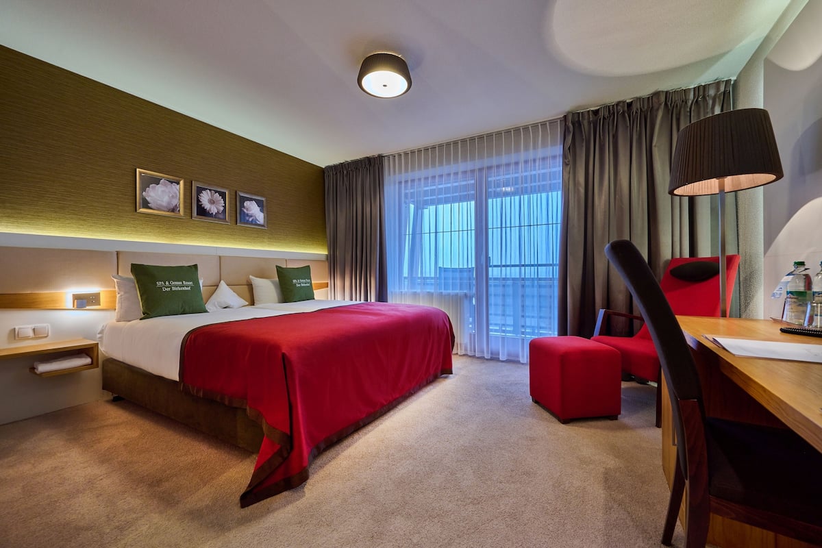 A hotel room with a large bed, a desk with a chair, a window with curtains, and a wall-mounted lamp.
