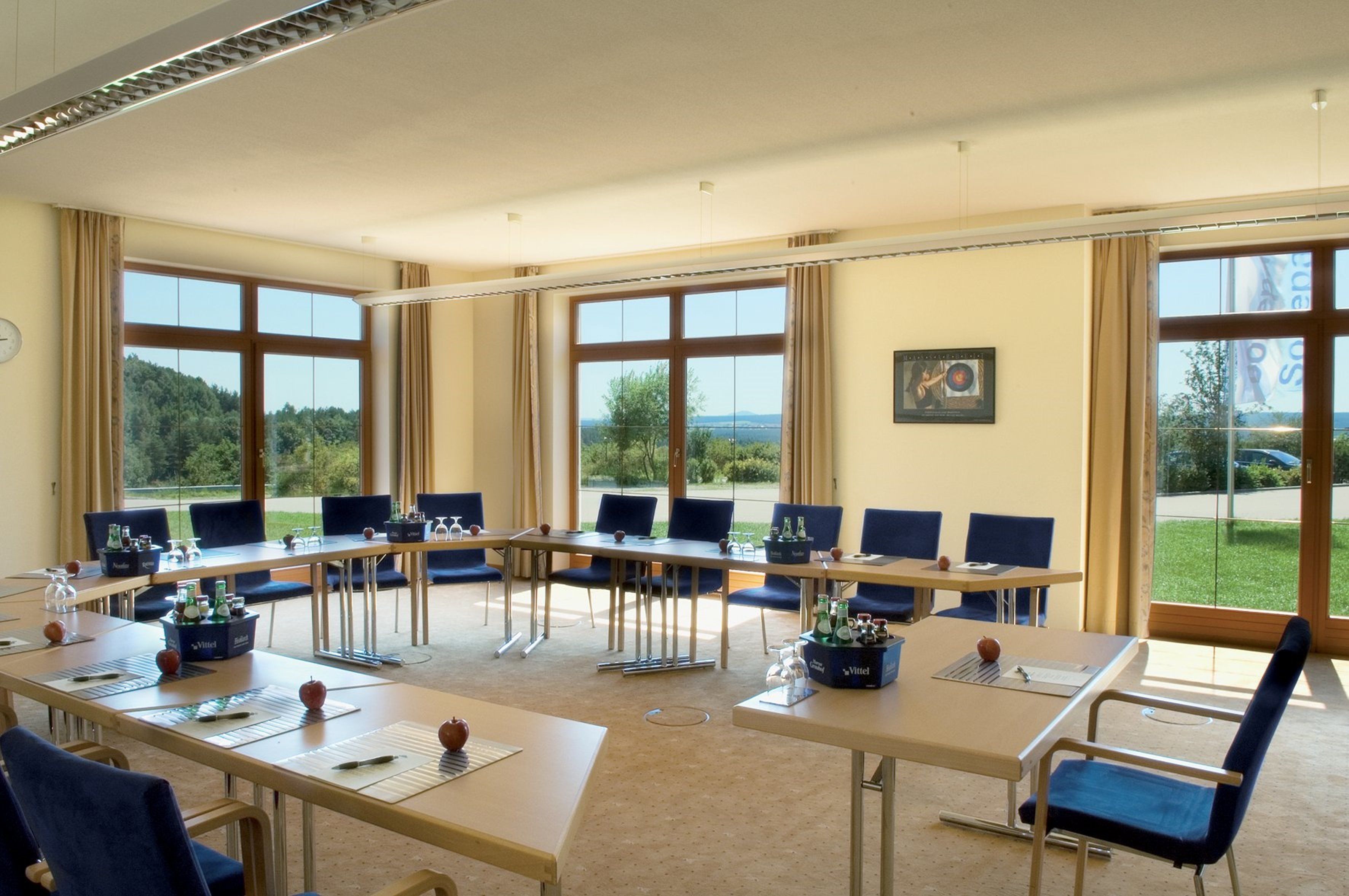 Un salón de conferencias con varias mesas y sillas, ventanas amplias que dan a zonas verdes, y un reloj en la pared.
