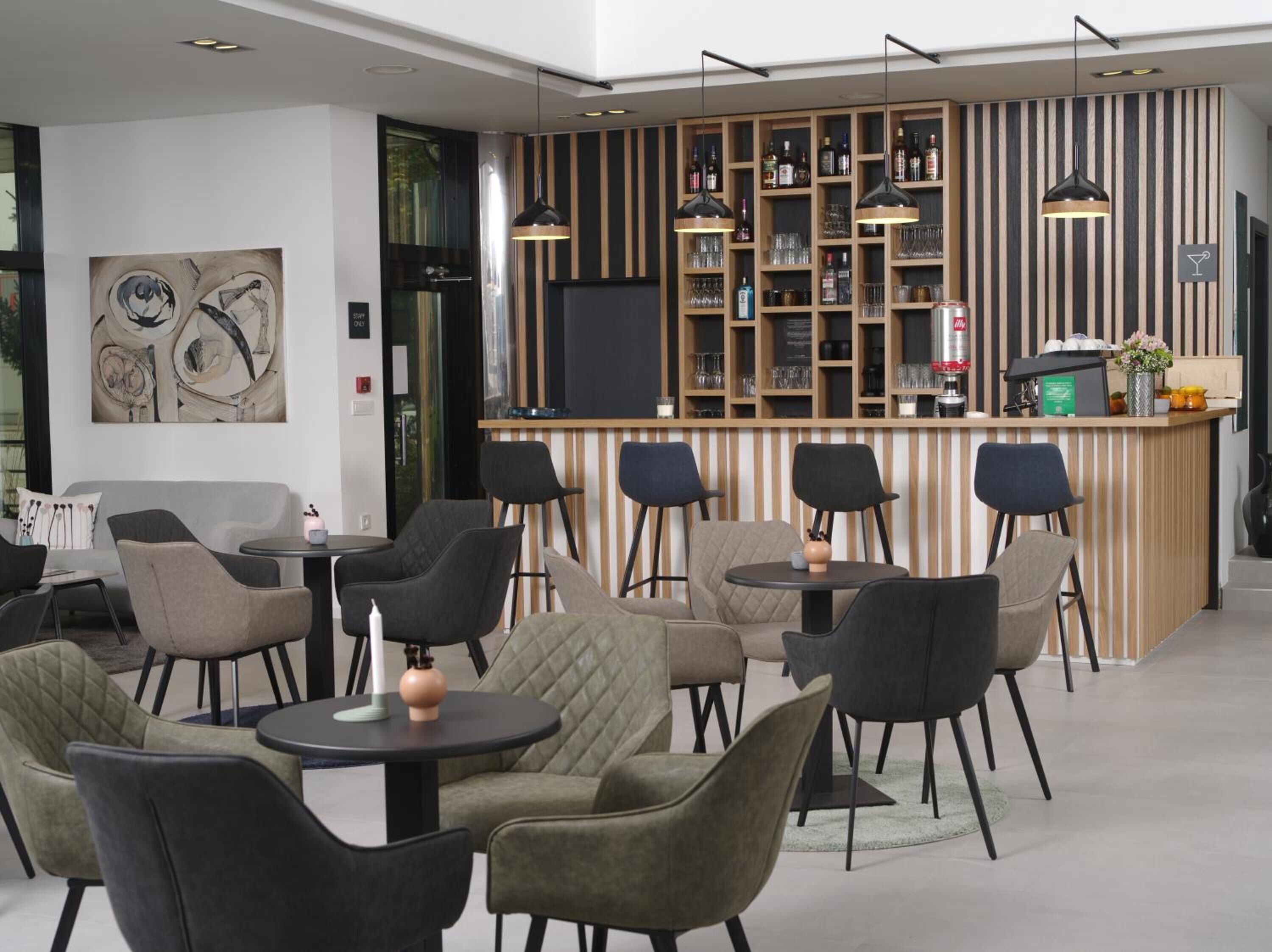 A modern bar area with a wooden counter, multiple seating options including round tables and chairs, and a large abstract painting on the wall.