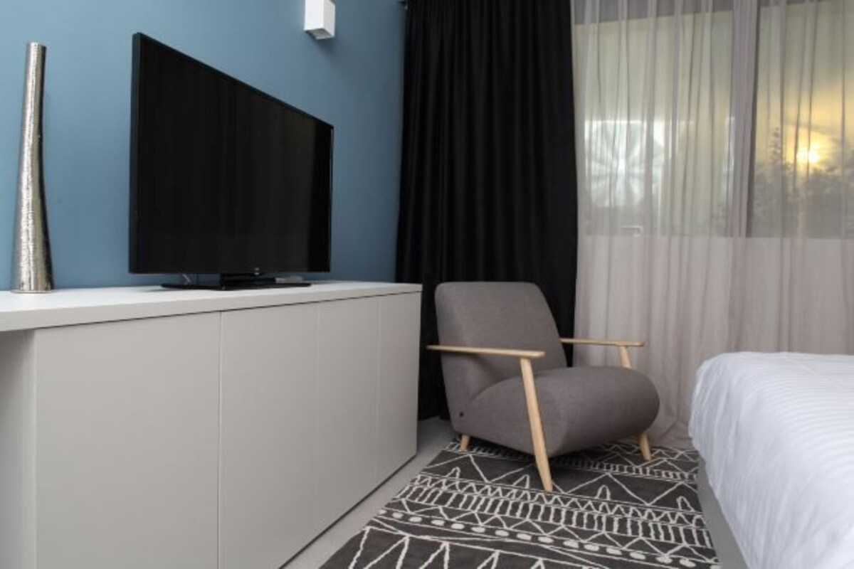 Ett modernt hotellrum med en platt-TV som är monterad på väggen, en grå klädd fåtölj, en vit säng och en mönstrad matta.