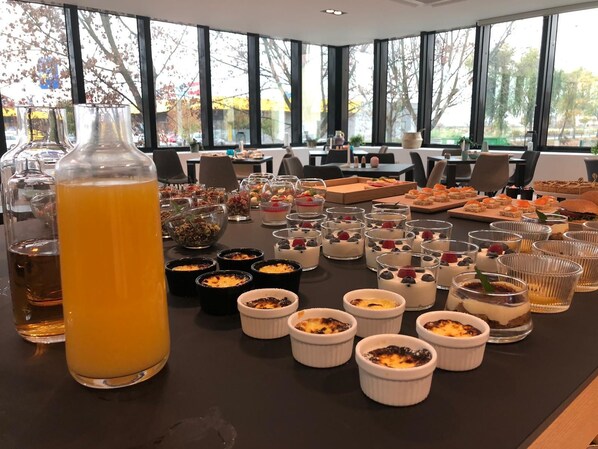 Una mesa de buffet con diversos postres y bebidas, ubicada en un salón con amplias ventanas y vistas a árboles.