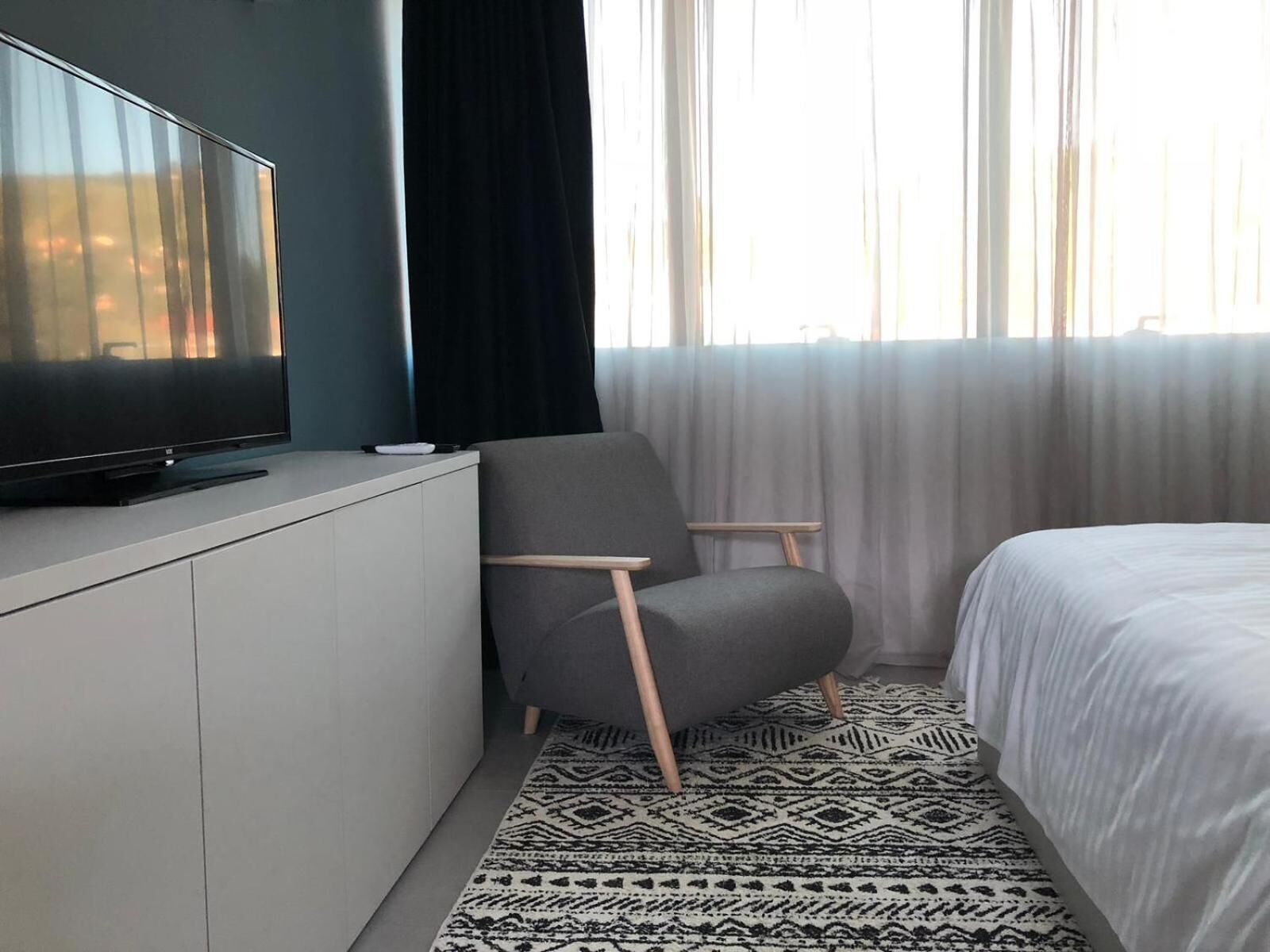 Quarto de hotel moderno com televisão de ecrã plano sobre um móvel branco, uma poltrona cinzenta, uma cama com roupa de cama branca e um tapete estampado.