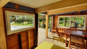 Una habitación con detalles de madera, un ventanal con vistas a un jardín con sillones de descanso y un comedor con mesa y sillas.