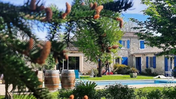Un edificio de piedra con contraventanas azules, una piscina y barriles de madera al frente.