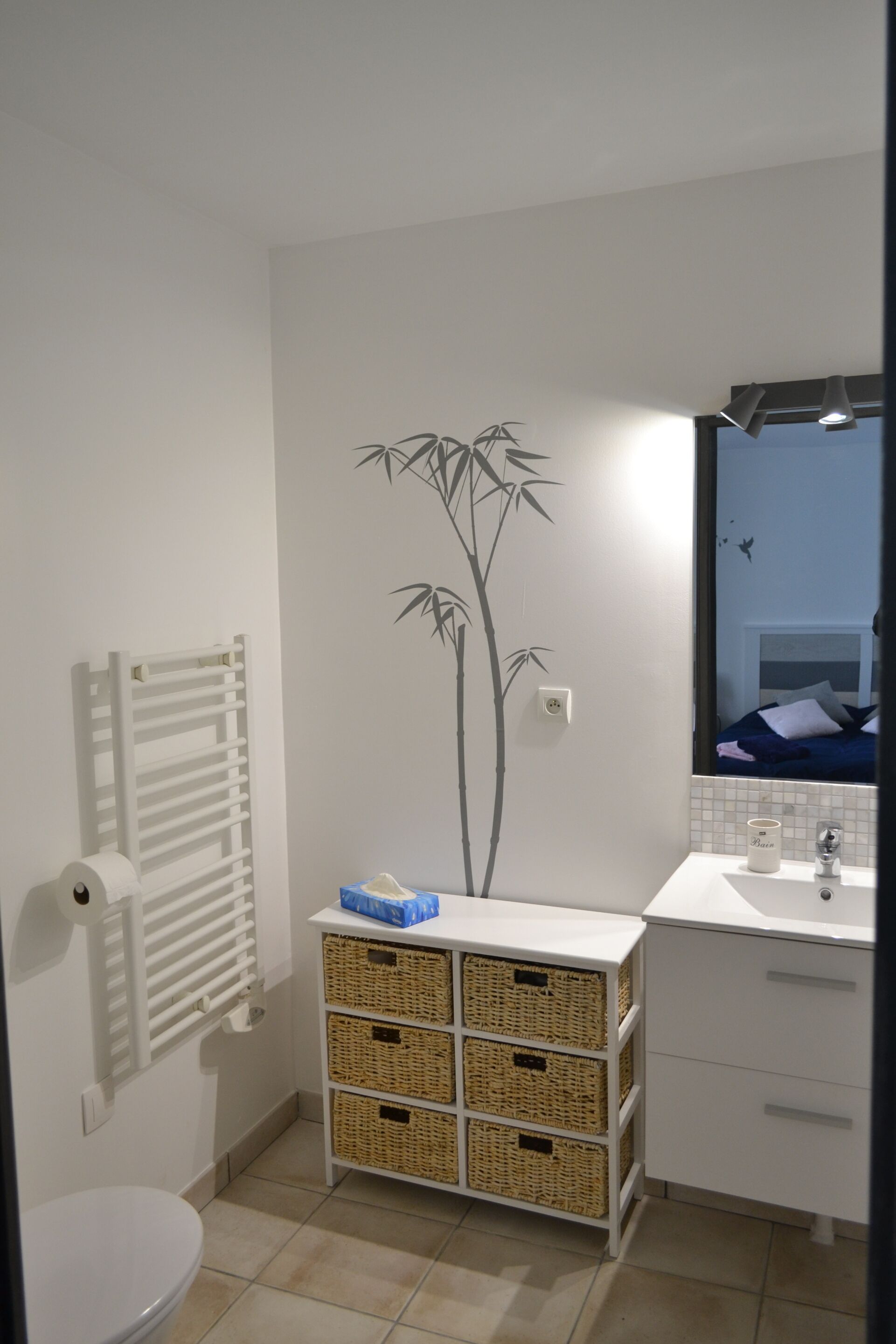 A bathroom with a toilet, a white sink, a mirror, and a wall-mounted radiator. There are wicker baskets on a shelf and a bamboo wall decal.