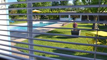 Udsigt gennem skodder til en baghave med swimmingpool, en gul parasol og havemøbler.