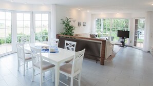 Uma sala de estar ampla com uma mesa de jantar branca e cadeiras, uma janela grande com vista para vegetação, e um espaço de estar aconchegante com um radiador e uma televisão.