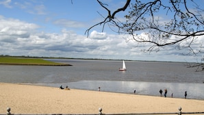 Uma praia arenosa com águas calmas, um veleiro e pessoas na orla.