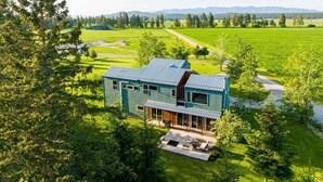 Ein modernes, zweistöckiges Haus mit einer großen Terrasse und einer überdachten Außenbereits, umgeben von üppiger Vegetation und ländlicher Landschaft.