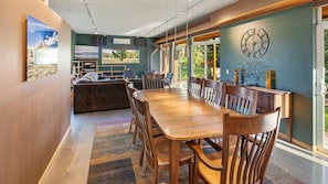 A modern dining area with a long wooden table and chairs, a large clock on the wall, and a view of the outdoors through large windows.