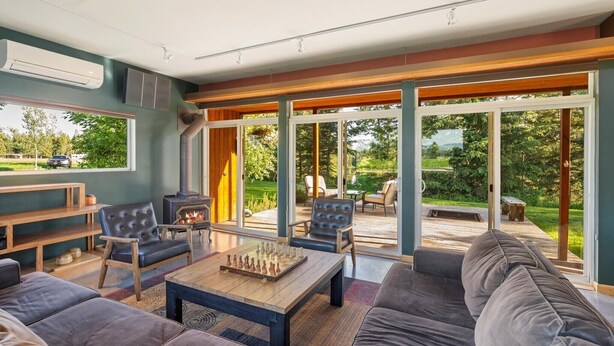 Ein geräumiges Wohnzimmer mit einer großen Couch, einem Couchtisch und einem Kamin. Es gibt mehrere Stühle und einen Esstisch. Große Glasschübe führen zu einer Außenpatio-Terrasse.